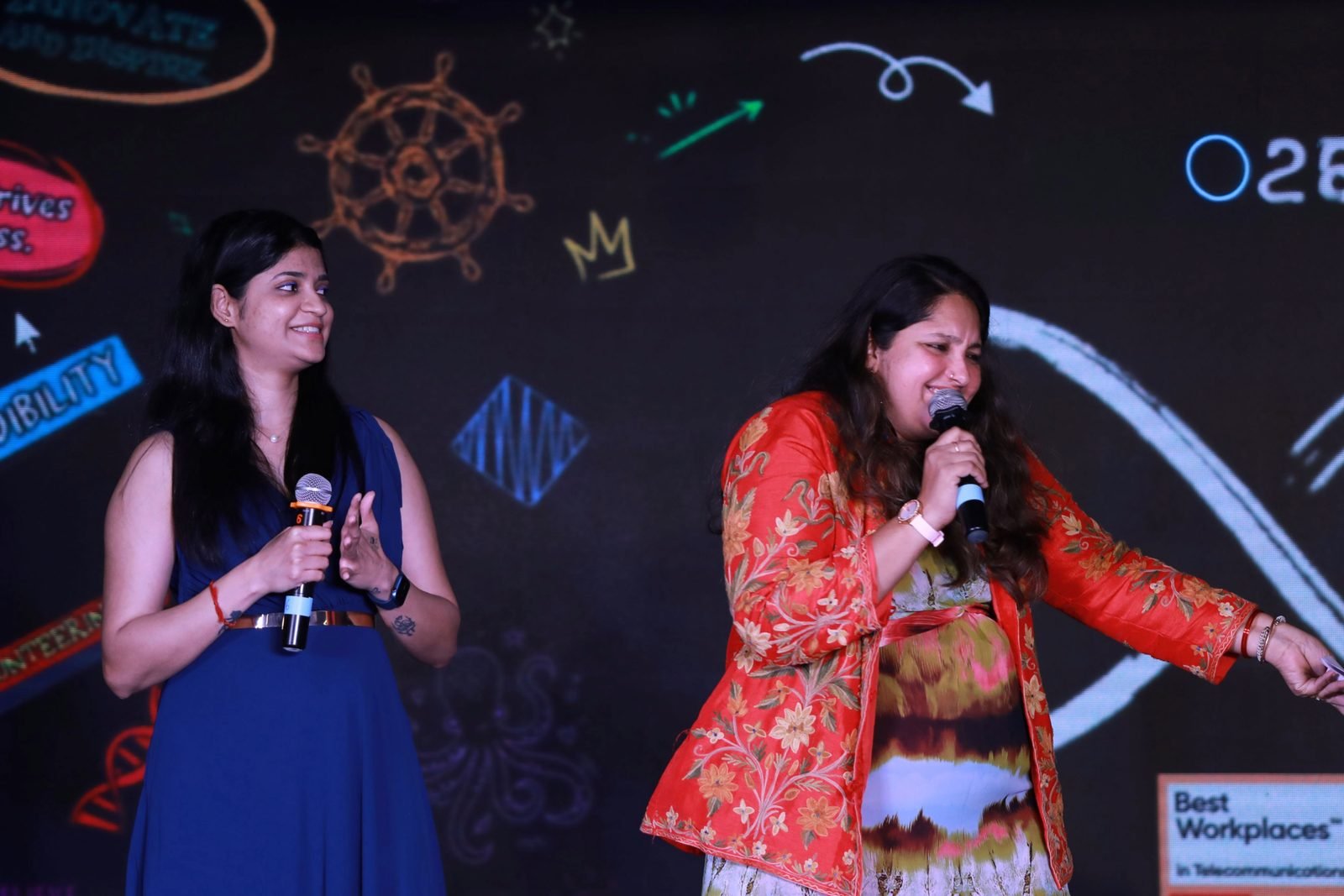 Pooja Aravalapalli co-hosting a workplace recognition night on stage, sharing the mic with a second host in front of a lit graphic backdrop
