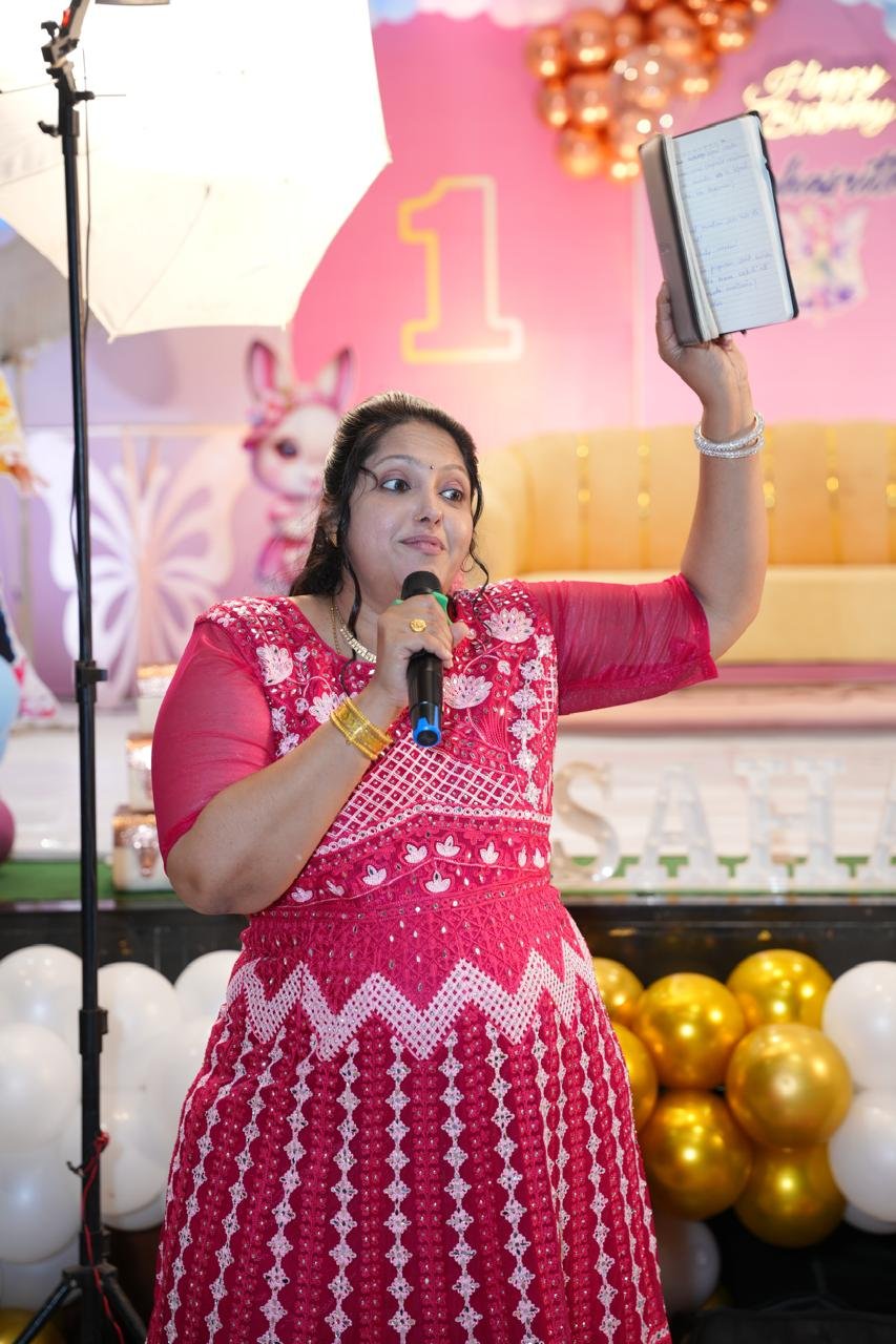 Pooja Aravalapalli hosting a first-birthday celebration, script in one hand and microphone in the other