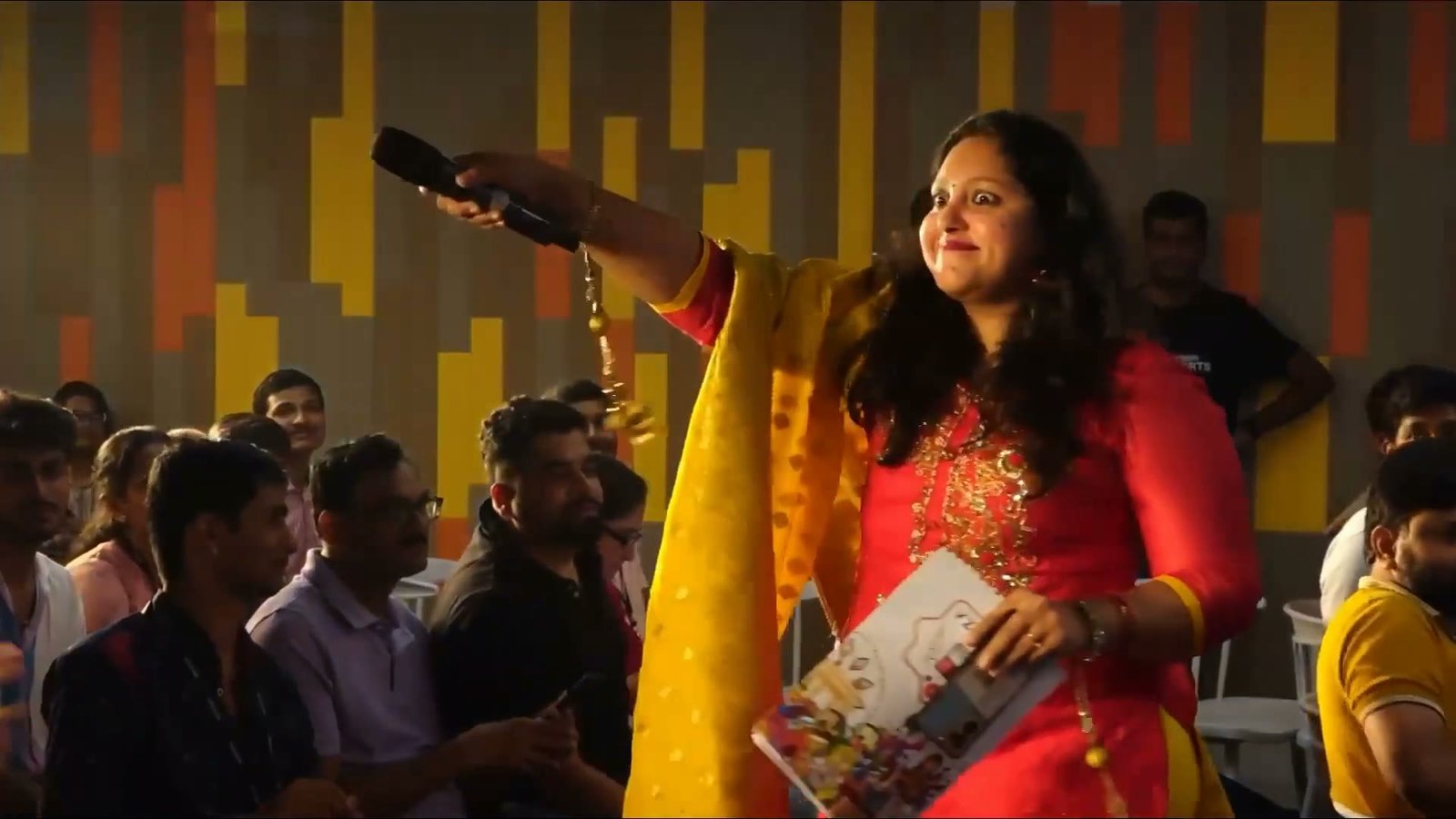 Pooja Aravalapalli holding a microphone out to a seated corporate audience, an attendee returning a thumbs-up