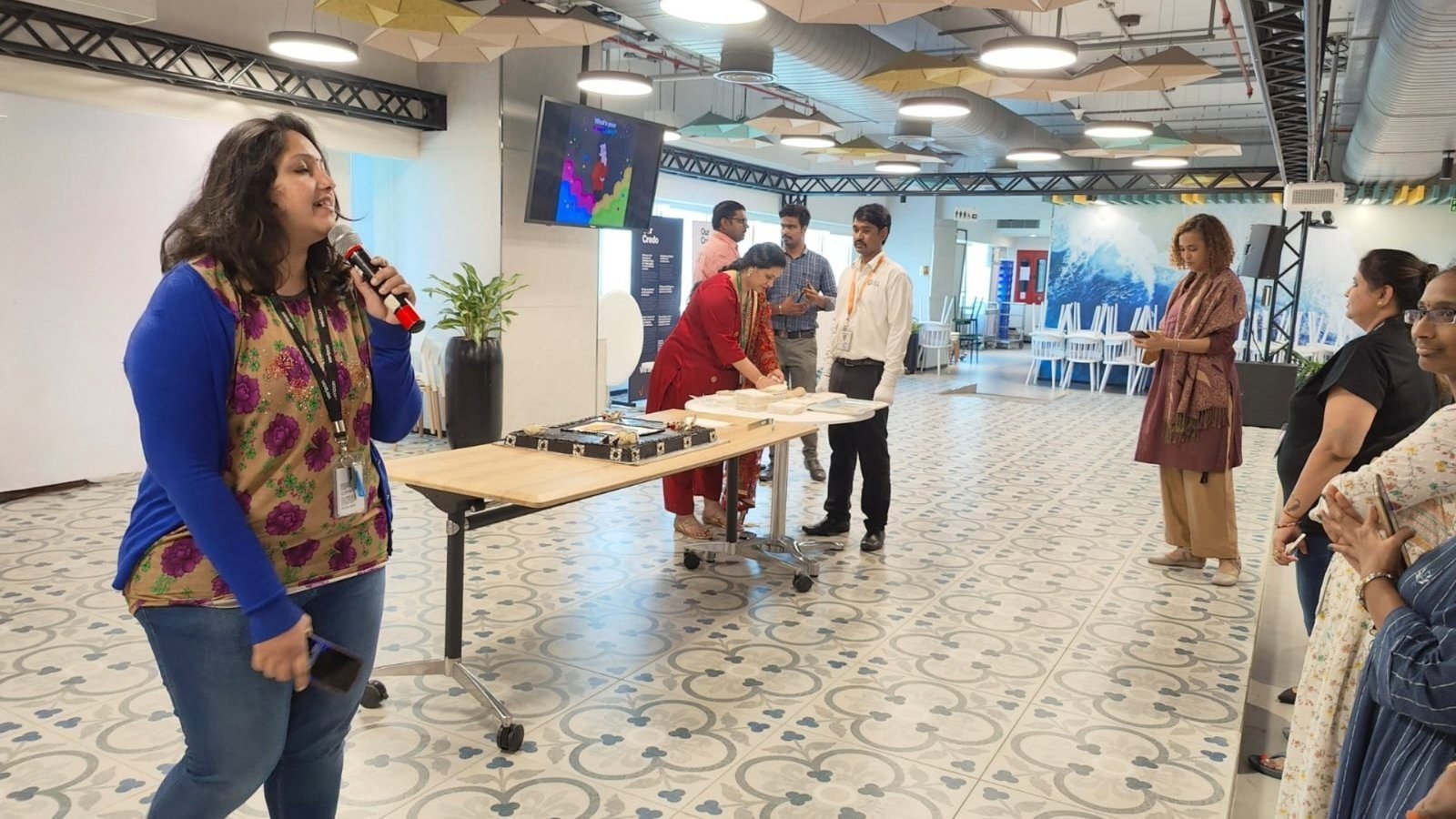 Pooja Aravalapalli hosting an in-office celebration, microphone in hand, colleagues gathered around a cake table on the floor behind her