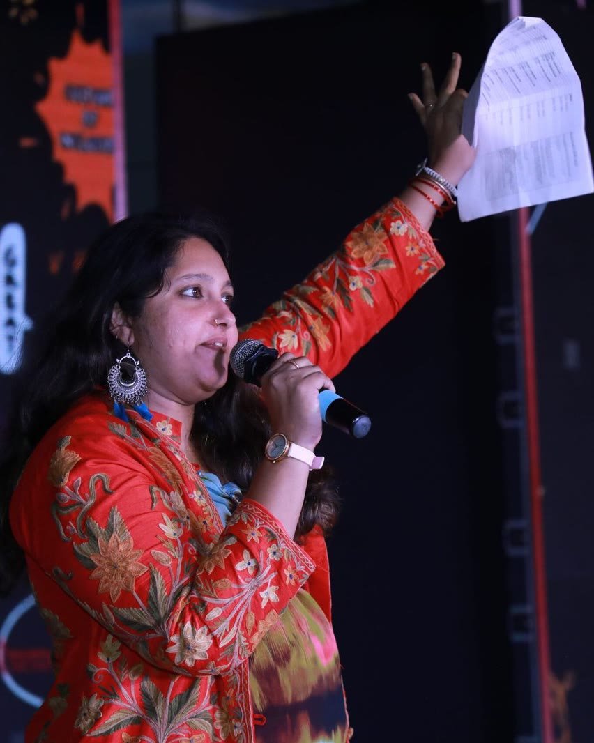 Pooja Aravalapalli hosting on a darkened stage, microphone in one hand and a run sheet raised overhead in the other