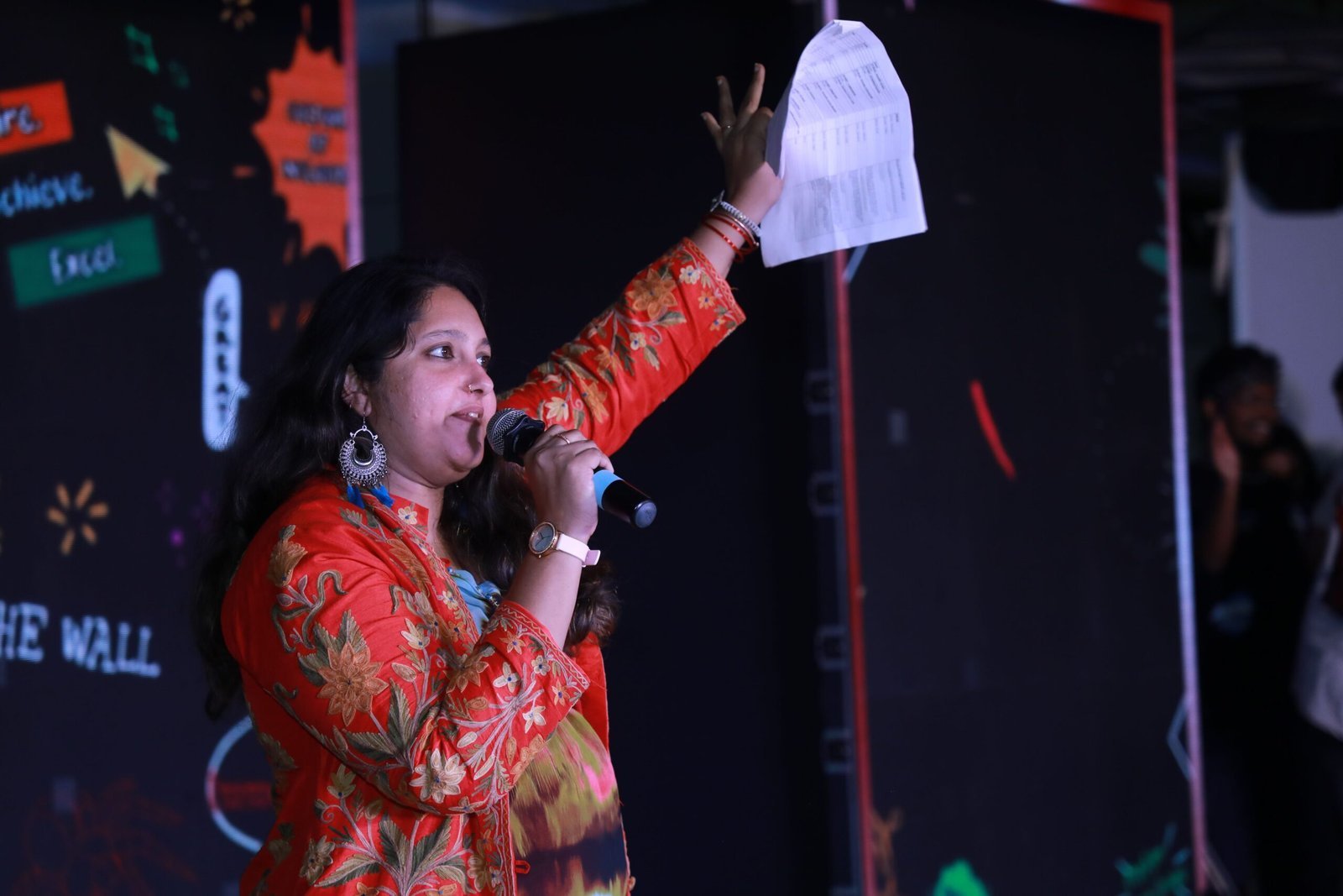 Pooja Aravalapalli on stage at a company town hall, run sheet held aloft, the organisation's values wall lit behind her