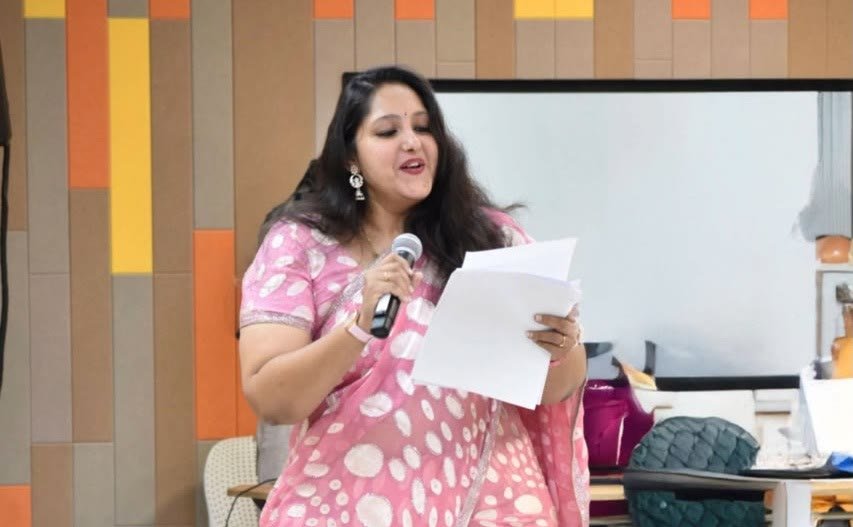Pooja Aravalapalli hosting indoors in a pink saree, microphone in one hand and a run sheet in the other, against a colourful panelled wall